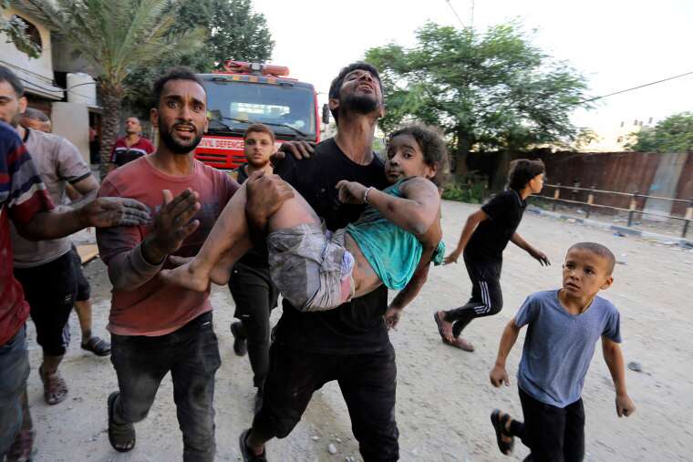 A man, surrounded by other people, carries a girl who is covered in dirt from building rubble along a road