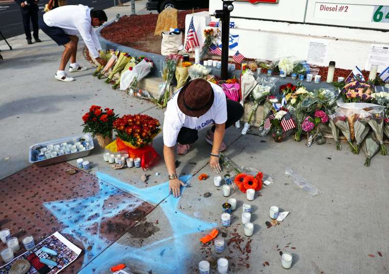 A person draws a Star of David on the side walk in front of a gas station among candles and flowers.